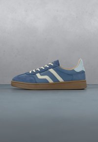 Baskets basses bleues avec des bandes blanches, des lacets blancs, un talon bleu clair et une semelle marron texturée sur une surface grise.