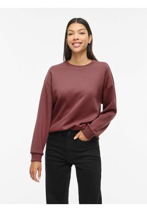 Noor naine pikkade tumedate juustega naeratab, kandes lahtist tumepunast pusa ja musta kõrge vöökohaga pükse tavalise valge tausta ees.