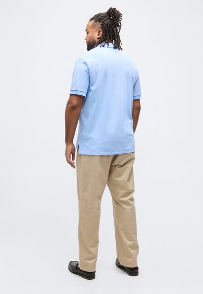 Άνδρας με dreadlocks που φοράει ανοιχτό μπλε πόλο πουκάμισο, μπεζ παντελόνι και μαύρα παπούτσια, στέκεται και κοιτάζει ελαφρώς προς τα αριστερά με φόντο ένα λευκό υπόβαθρο.