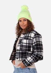 Cropped black and white checked flannel jacket with a single chest pocket, worn over a beige sweater and paired with a bright green knit beanie.