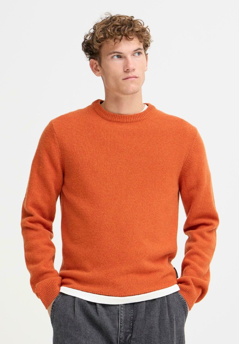 Oransje strikket genser med rund hals, lange ermer og ribbede mansjetter. Brukt over en hvit skjorte, med myk tekstur og middels passform.