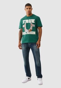 Zielony bawełniany T-shirt z napisem "TRUE RELIGION" w białym kolorze oraz szczegółowym logo. Do kompletu niebieskie dżinsy i białe trampki.
