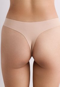 Frau trägt nahtlose hautfarbene Tanga-Unterwäsche, zeigt den unteren Rücken und die oberen Oberschenkel vor einem schlichten Hintergrund.