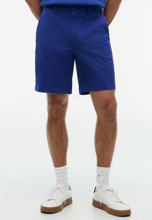 Mann, der blaue, maßgeschneiderte Shorts, weiße Socken und weiße Sneaker trägt, steht vor einem schlichten hellen Hintergrund.