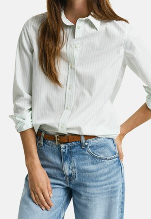 Femme portant une chemise blanche à manches retroussées rentrée dans un jean bleu taille haute avec une ceinture marron, la main posée sur la hanche.