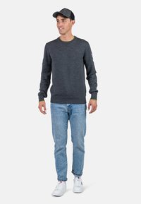 Grauer Strickpullover mit rundem Ausschnitt, gelben Akzenten am Kragen, kombiniert mit hellblauen Jeans und weißen Sneakers. Graue Mütze inklusive.