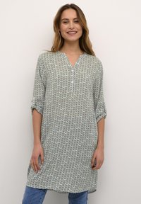 Tunique légère et verte avec un motif en zigzag, devant boutonné, poche poitrine et manches retroussées. Associée à un jean bleu pour un look décontracté.