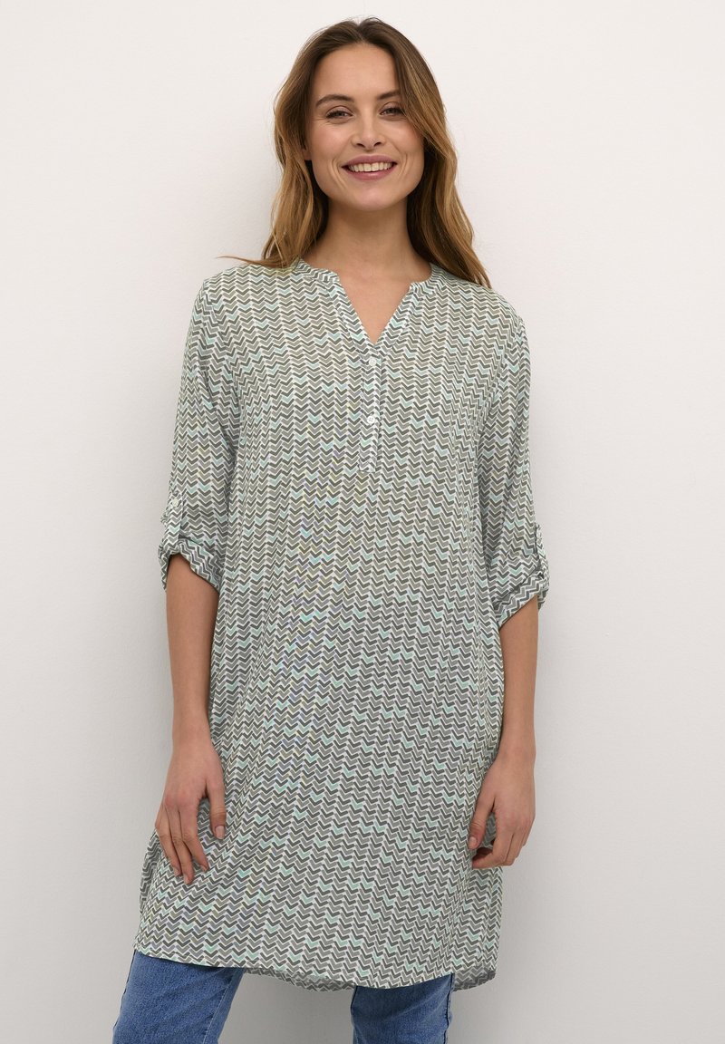 Tunique légère et verte avec un motif en zigzag, devant boutonné, poche poitrine et manches retroussées. Associée à un jean bleu pour un look décontracté.