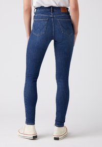 Jeans ajustados azules de mezclilla, con una cinturilla alta, bolsillos traseros con costuras sutiles, y combinados con zapatillas de lona blancas.