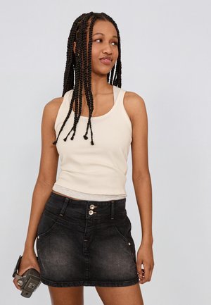 Tânără cu cozi lungi împletite, purtând un top alb fără mâneci și o fustă din denim negru, ținând o cameră vintage în mâna dreaptă.