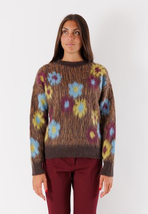Donna con lunghi capelli scuri che indossa un maglione marrone peloso con motivi floreali blu, gialli e rossi e pantaloni bordeaux, rivolta in avanti.