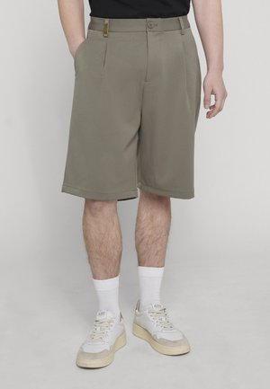 Dolna część ciała osoby ubranej w beżowe krótkie spodenki do kolan, białe wysokie skarpety i białe trampki, stojącej z jedną ręką w kieszeni.