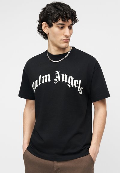 Νεαρός άνδρας με σκούρα σγουρά μαλλιά κοιτάζει δεξιά, φοράει μαύρο μπλουζάκι "Palm Angels", ασημένιο κολιέ αλυσίδα και καφέ παντελόνι με τα χέρια στις τσέπες.