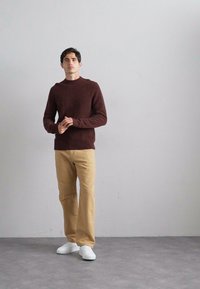Maglione marrone a maglia, pantaloni beige e sneakers bianche. Il maglione ha un collo rotondo a coste. L'outfit presenta una vestibilità comoda e uno stile casual.