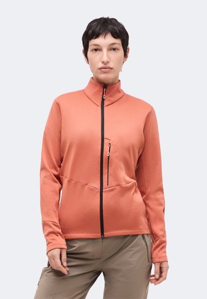 Asmuo dėvi koralo spalvos užsegamą striukę su juodu užtrauktuku ir užtrauktuku krūtinės kišenėje, derinama su smėlio spalvos kelnėmis ant paprasto fono.