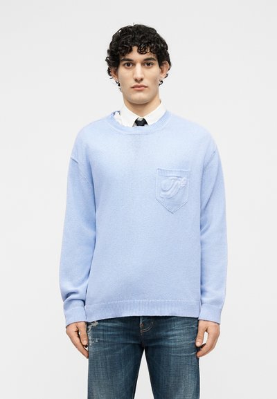 Jeune homme aux cheveux bouclés foncés portant un pull bleu clair par-dessus une chemise blanche et une cravate noire, assorti à un jean bleu.