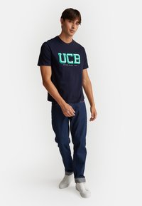 Námořnická modrá bavlněná tričko s velkým tyrkysovým logem "UCB" a textem "ZALOŽENO 1965". V kombinaci s tmavě modrými džíny a bílými teniskami.