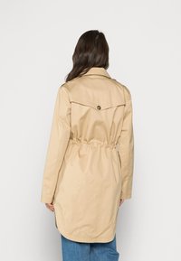 Trench beige con cintura ceñida, con hombreras y dobladillo redondeado. Tejido ligero, mangas largas y un detalle de botón en la parte posterior.