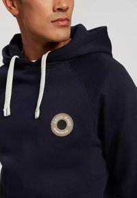 Granatowa bluza z kapturem o teksturowanym wykończeniu, z kieszenią kangurzą, białymi sznurkami i okrągłą łatą z logo na klatce piersiowej.