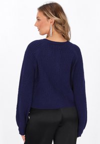 Donna vista di profilo che indossa un maglione a maglia testurizzata blu navy, lunghi orecchini a nastro argentati e pantaloni neri su uno sfondo bianco.