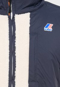Marineblå jakke med en kremhvit imitasjonsull foran, med glidelåslukking og K-WAY-logo på brystet. Teksturt fleece kontrasterer med glatt stoff.