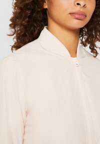 Femeie cu păr creț care poartă o jachetă bej deschis cu fermoar și guler înalt, prezentată de la nivelul umerilor în sus pe un fundal simplu.