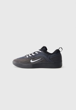 Sort og mørkegrå lavtop atletiksneaker med hvide snørebånd og et hvidt Nike swoosh-logo på siden, vist i profil mod en grå baggrund.