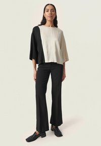 Haut noir et crème en bloc de couleurs, avec des manches larges, assorti à un pantalon noir ajusté et des chaussures noires à enfiler sur un fond neutre.