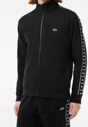 Mężczyzna ubrany w czarną sportową kurtkę na zamek i spodnie z białymi paskami z logo Lacoste na rękawach i nogawkach, stoi na białym tle.
