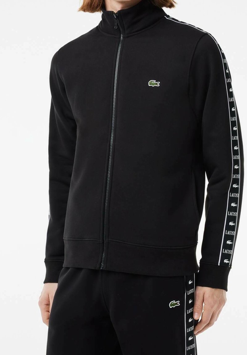 Homme portant une veste de sport noire zippée et un pantalon avec des bandes blanches avec logo Lacoste sur les manches et les jambes, debout devant un fond blanc.
