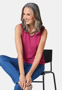 Magenta mouwloze top met een ronde halslijn, gecombineerd met blauwe jeans. Model zit in een ontspannen houding en toont details van de textuur van de stof.