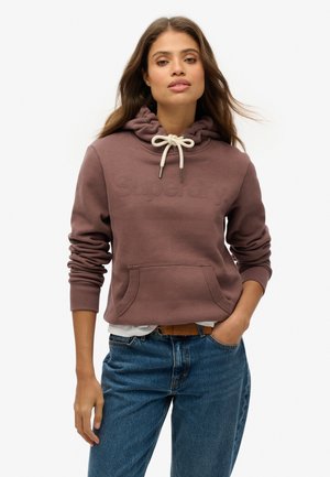Donna con lunghi capelli che indossa un hoodie malva con tasca frontale e logo "Superdry", abbinato a jeans blu e una cintura marrone, in piedi su uno sfondo bianco.