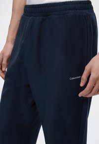 Μπλε σκούρο παντελόνι φόρμας Calvin Klein με ελαστική μέση και πλαϊνές τσέπες, φορεμένο από ένα άτομο με τα χέρια χαλαρά στα πλευρά.