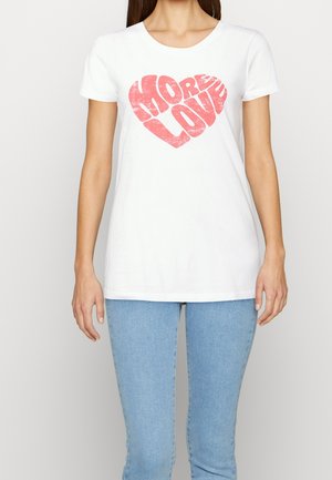 Kvinde iført hvid T-shirt med rød "MORE LOVE" tekst formet som et hjerte, kombineret med lyseblå jeans, mod en ensfarvet baggrund.