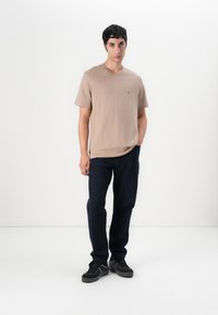 T-shirt beige à col en V en tissu doux, associé à un jean bleu foncé. Le modèle porte des baskets noires, posé contre un fond blanc uni.