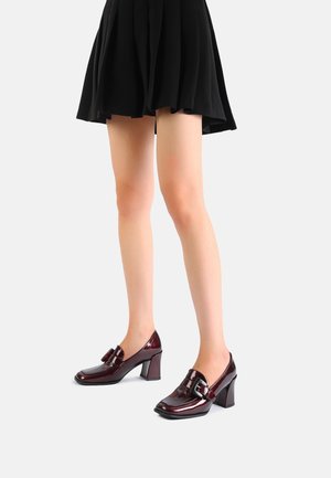 Personne portant des chaussures à talons carrés rouge foncé brillantes avec des boucles et une jupe noire plissée, debout sur un fond clair.