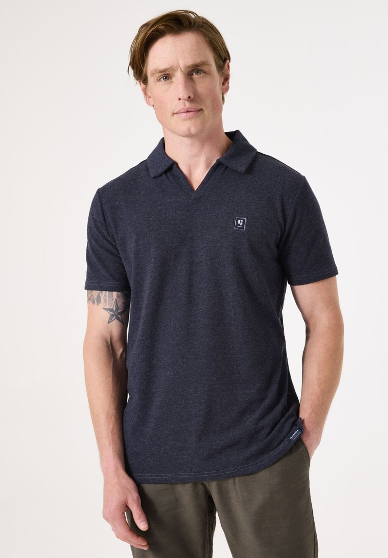 Marineblauwe poloshirt met korte mouwen en een V-hals ontwerp. De stof heeft een zachte textuur. Klein logo op de linkerkant van de borst.