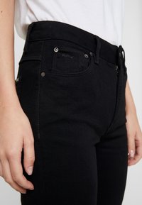 Jeans de mezclilla negra con ajuste slim, que incluyen bolsillos frontales, detalles metálicos y un sutil logo en el bolsillo.