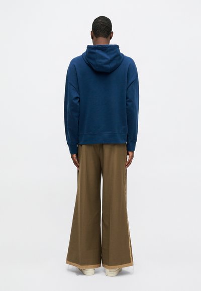 Sudadera con capucha de color azul marino con puños de canalé, combinada con pantalones anchos marrones que tienen un ribete en contraste en el dobladillo y zapatillas blancas.