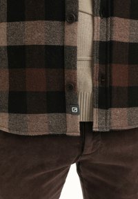 Bruin en zwart geruit flanellen shirt met donkere knopen en een subtiel logo, gelaagd over een lichtbeige gebreid truitje, gecombineerd met donkerbruine corduroy broek.