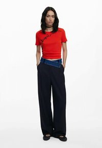 Czerwona dopasowana koszulka z nadrukiem; w zestawieniu z granatowymi szerokimi spodniami z paskiem w stylu dżinsowym. Modelka stoi z rękami w kieszeniach.