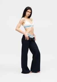 Γυναίκα που φοράει λευκό σουτιέν-μπραλέτα και εσώρουχα με μπλε σχέδιο, κατεβάζοντας φαρδιά, σκούρα μπλε τζιν με άνετη εφαρμογή, όρθια και ξυπόλυτη.