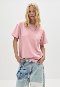 Ung kvinde med løst blonde h�år iført en pink graphic t-shirt og patchwork denim jeans, mens hun holder en blå denim håndtaske.