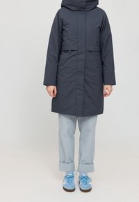 Manteau matelassé gris foncé avec capuche, manches longues et ceinture à la taille. Porté par-dessus un pantalon bleu clair retroussé et des baskets bleues.