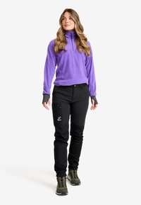 Kvinna står klädd i lila fleecepullover, svarta cargobyxor med logga och gröna vandringsskor, och ser lätt åt sidan.