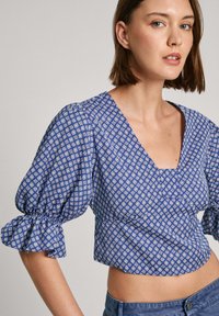 Modrá vzorovaná crop top s V-výstrihom, nadýchanými krátkymi rukávmi a volánovými manžetami. Tkanina má geometrický vzor v bielej farbe.