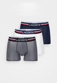 Trois paires de boxers : une rayée en gris et noir, une unie en bleu marine, et une unie en blanc, toutes avec une ceinture élastique de marque.