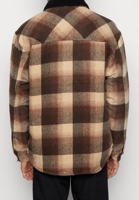 Camicia a quadri realizzata in un morbido tessuto testurizzato marrone e beige, con colletto nero e orlo arrotondato, cuciture e polsini posteriori visibili.