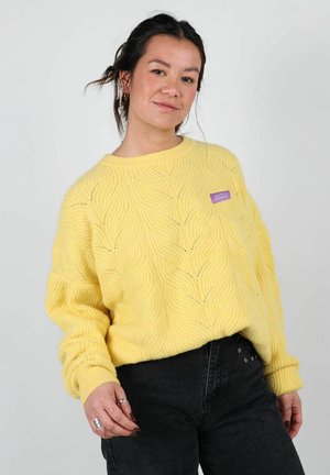 Pull en tricot jaune avec un motif en vagues, col rond, et poignets côtelés. Présente une petite étiquette violette sur le côté gauche. Associe à un pantalon noir.
