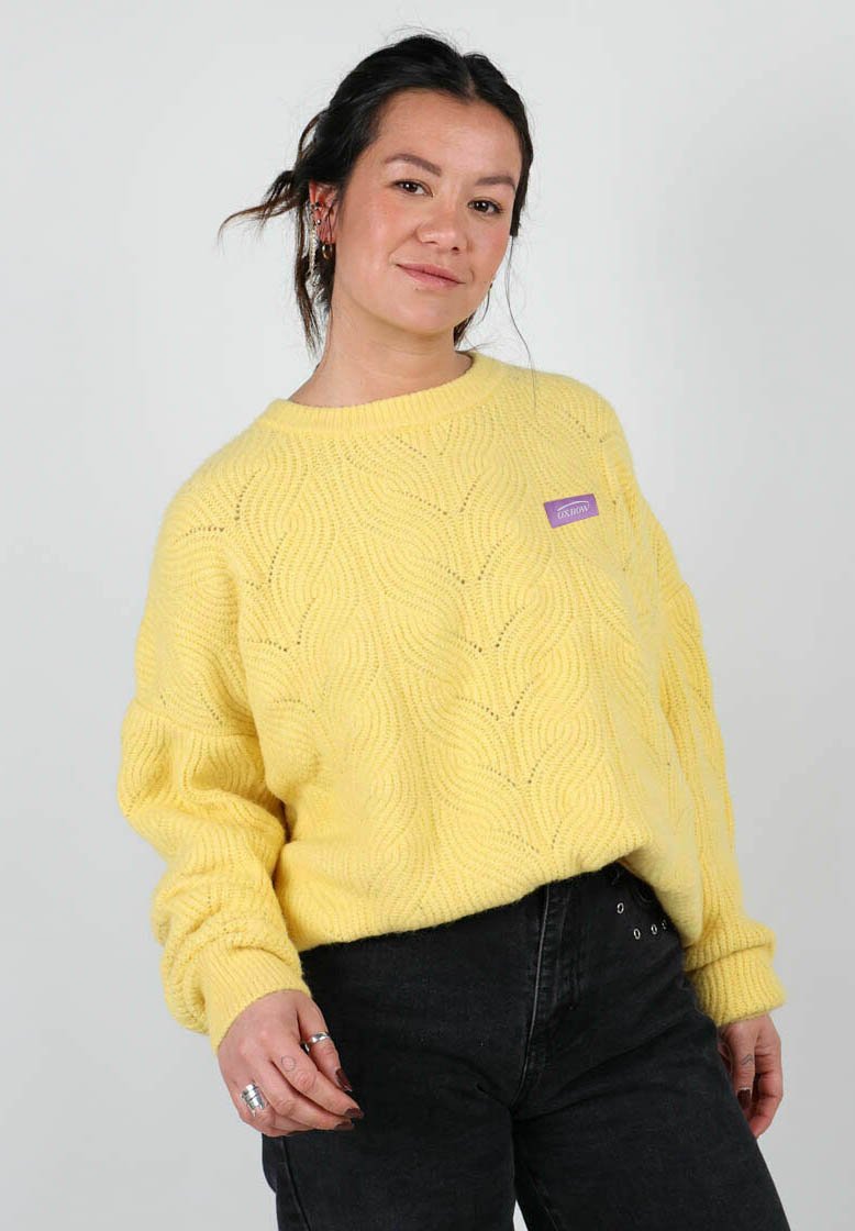 Pull en tricot jaune avec un motif en vagues, col rond, et poignets côtelés. Présente une petite étiquette violette sur le côté gauche. Associe à un pantalon noir.